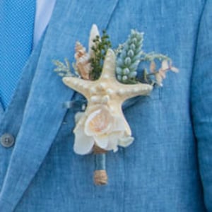 Op de afbeelding: Een beige zeester met een witte schelp en blauwe en groene bloemmotieven, een bruiloft corsage.