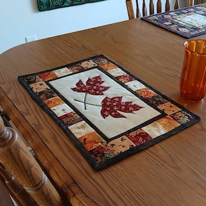 Może przedstawiać: Prostokątny bieżnik z kremowym środkiem z dwoma czerwonymi liśćmi klonu. Obramowanie to patchwork z tkanin o tematyce jesiennej. Pomarańczowy szklany wazon stoi na drewnianym stole.