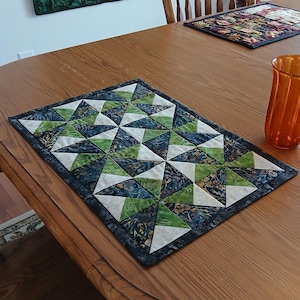 Puede incluir: Camino de mesa acolchado hecho a mano con un patrón geométrico de estrellas en tonos verdes, azules y blancos. El camino de mesa es rectangular y está sobre una mesa de madera, junto a un vaso naranja y otros objetos decorativos.