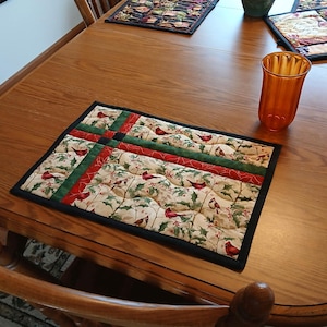 Op de afbeelding: Een rechthoekige, doorgestikte placemat met een crèmekleurige achtergrond en een patroon van rode kardinalen, groene hulstbladeren en rode bessen. De placemat heeft een zwarte rand en rode en groene stroken in een kruisvorm.