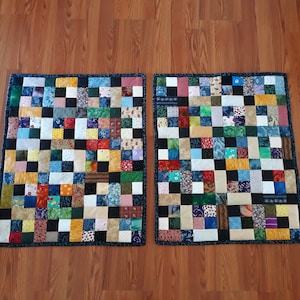 May include: Two rectangular quilts featuring a patchwork design composed of numerous small, multi-coloured fabric squares in various patterns, including solids, florals, and geometric prints, all bordered by a dark blue fabric with a subtle pattern.
