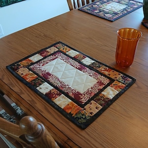 May include: A quilted table runner with a central cream-colored diamond pattern, framed by a border of autumn-themed fabrics in shades of orange, burgundy, and green. An amber-colored glass sits on the wooden table.