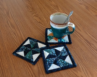 May include: A teal mug with a silver spoon rests on a quilted coaster with a geometric design. Two additional coasters with similar patterns are visible. The coasters feature white, teal, and green fabrics with a dark blue border. The items are on a wooden table.