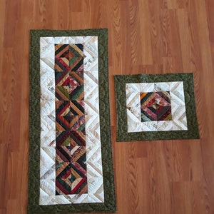 May include: Two quilted table runners with a green border and a white background. The larger runner has a geometric pattern in shades of brown, red, and green. The smaller runner has a similar pattern but is smaller in size.