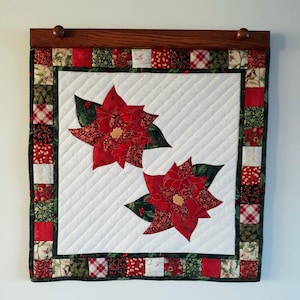 以下が含まれることがあります： 白い背景と赤と緑の縁取りのあるキルトの壁掛け。壁掛けの中央には、緑の葉が付いた赤いポインセチアの花が2つあります。縁取りは、さまざまな色と模様の小さな布の正方形でできています。