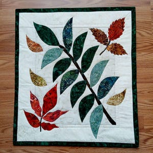 May include: A square quilted wall hanging with a central branch and green and blue leaves. Red, brown, and yellow leaves are scattered around. The background is white with a green border, creating a fall-themed design.