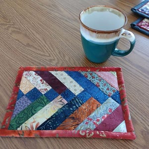May include: A colorful patchwork fabric coaster with a red border. The coaster is made of various fabrics with different patterns and colors, including blue, green, purple, and orange. A teal mug is sitting on a wooden table next to the coaster.