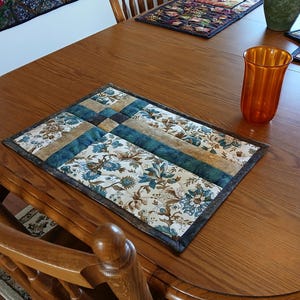 Peut inclure: Un set de table rectangulaire matelassé présentant un motif en patchwork avec des bandes alternées de tissu floral dans des tons bleu, crème et marron, bordé d'une bordure en tissu marron foncé. Le set est cousu avec un motif en grille.