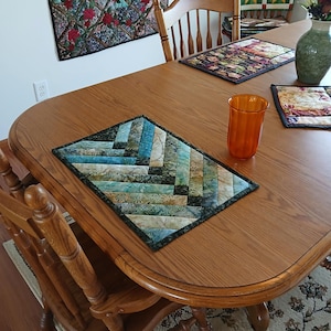 Pode incluir: Um lugar americano acolchoado retangular com um padrão de espinha de peixe em tons de azul, verde, turquesa e bege. O tecido apresenta uma estampa botânica sutil e é finalizado com uma borda verde escura ao longo das extremidades da peça.