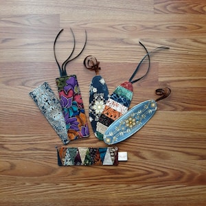 Op de afbeelding: Een set van zes kleurrijke stoffen bladwijzers met verschillende patronen en ontwerpen. De bladwijzers zijn gemaakt van stofresten en hebben een verscheidenheid aan kleuren, waaronder blauw, groen, rood en geel. De bladwijzers zijn aan elkaar gebonden met een zwart lint.