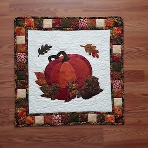 Op de afbeelding: Een gequilt wandkleed met een witte achtergrond en een bruine, oranje en rode pompoen omringd door herfstbladeren. De rand is gemaakt van patchwork stof in tinten bruin, oranje, rood en groen.