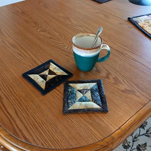 May include: Two square fabric coasters featuring a geometric patchwork design in shades of brown, tan, and teal, positioned next to a two-toned green and white ceramic mug containing a silver spoon, all resting on a wooden surface.