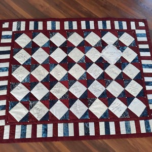 Puede incluir: Un edredón hecho a mano con un patrón de diamantes en rojo, blanco y azul. El edredón presenta mensajes escritos a mano en algunos de los diamantes blancos. El borde tiene rayas alternas rojas, blancas y azules. Una pieza decorativa con texto está en el centro.