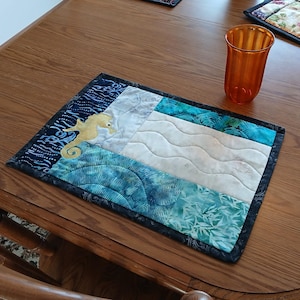 Könnte beinhalten: Ein rechteckiger, gesteppter Tischset mit einem Seepferdchen-Design. Das Tischset hat blaue, weiße und türkisfarbene Stoffabschnitte mit einer Applikation eines Seepferchens. Der Rand ist dunkelblau, auf einem Holztisch.