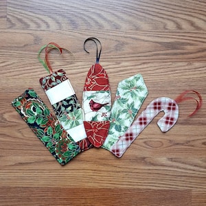 May include: A collection of handmade Christmas ornaments in various shapes. The ornaments feature festive patterns like holly, plaid, and floral designs, with red, green, and white colors. Each ornament has a ribbon for hanging.