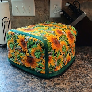 May include: A rectangular kitchen appliance cover featuring a vibrant sunflower pattern in yellow, orange, and green. The cover has a green border and is likely designed to protect a toaster or similar appliance. The floral design adds a touch of nature to the kitchen.