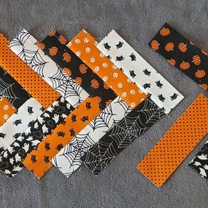 Pode incluir: Faixas de tecido com tema de Halloween com padr&otilde;es de ab&oacute;boras, teias de aranha, fantasmas e bolinhas nas cores laranja, preto e branco. As faixas est&atilde;o dispostas sobre uma superf&iacute;cie cinza, mostrando um tema festivo sazonal.