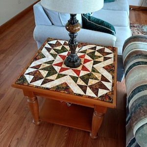 May include: A square wooden end table with a lower shelf, topped with a fabric quilt featuring a geometric star pattern in shades of red, green, brown, and cream. A table lamp with a dark, textured base and a light-coloured drum shade sits on top.