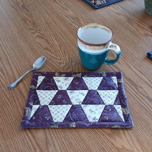May include: A handmade quilted coaster with a geometric pattern of purple and cream triangles. A teal mug with a brown rim sits nearby, along with a silver spoon. The coaster is on a wooden table.