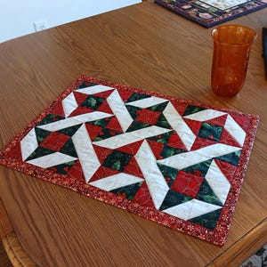 May include: A rectangular Christmas-themed quilted table runner. The runner features a pattern of white, red, and green geometric shapes. The border is red with a small floral design. The runner is on a wooden table.