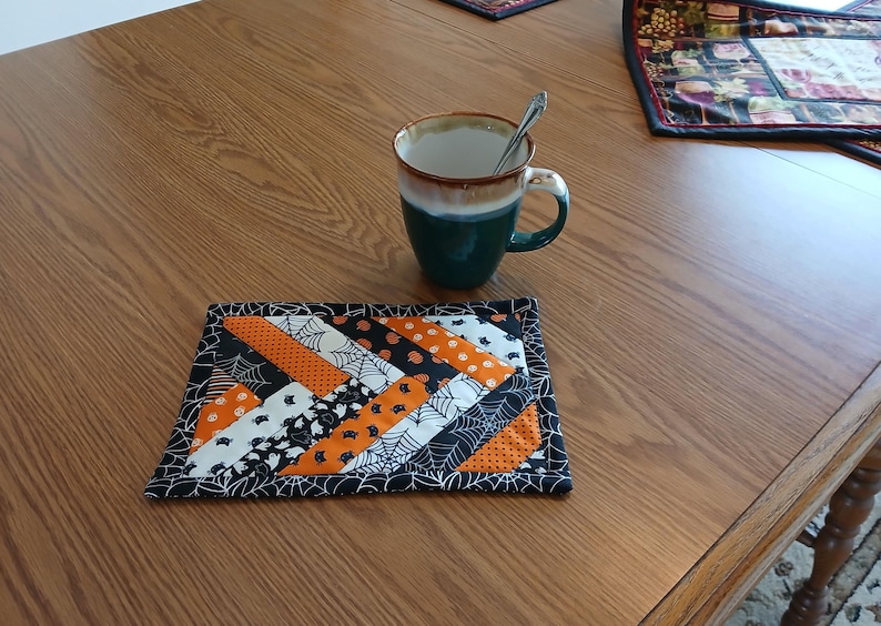 Pode incluir: Uma base de copo artesanal com tema de Halloween e design de patchwork. A base de copo apresenta tecidos laranja, preto e branco com padr&otilde;es de teias de aranha, morcegos e ab&oacute;boras. Uma caneca verde-azulado e castanha com uma colher est&aacute; na mesa.