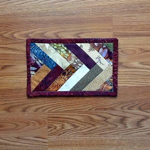 May include: A rectangular quilted fabric piece with a herringbone pattern. The design features various fabric strips in shades of blue, cream, brown, and purple, bordered by a deep burgundy fabric. The piece is likely a coaster or small mat.