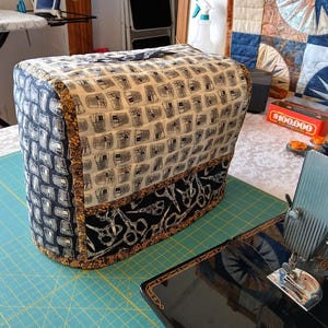 May include: A quilted sewing machine cover with a black, white, and beige pattern. The cover has a handle and is trimmed with a black fabric featuring a scissors design. The cover sits on a teal cutting mat.