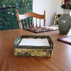 May include: A fabric napkin holder with a green grape pattern. The holder is filled with white napkins and sits on a wooden table. The trim is a dark green fabric. A vase and placemats are in the background.