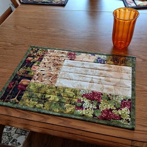 May include: A quilted placemat with a wine theme, featuring sections with grapes, wine bottles, and corks. The placemat is bordered with a green fabric and sits on a wooden table. An orange glass is on the table.