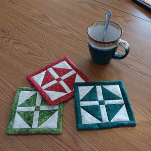 Könnte beinhalten: Drei quadratische, gesteppte Untersetzer in Rot, Grün und Türkis mit weißen geometrischen Designs. Eine Keramiktasse mit einem Löffel darin steht in der Nähe auf einem Holztisch. Die Untersetzer sind etwa 10 cm groß.