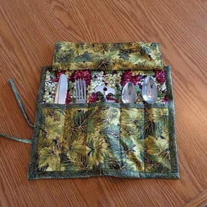 May include: A fabric cutlery roll with a green and gold leaf pattern, holding a knife, fork, spoon, and bottle opener. The roll is open, displaying the silverware. The fabric has a border of green fabric and ties to secure the roll.