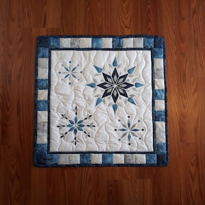 May include: A white quilted wall hanging with a blue and white border. The center of the hanging features five blue and white snowflake designs.
