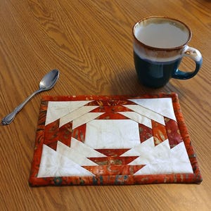 May include: A red, white, and green quilted coaster with a geometric pattern. The coaster is made of fabric and has a white center with red and green triangles surrounding it.