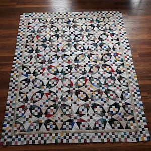 Op de afbeelding: Een patchworkdeken met een geometrisch ontwerp met verschillende kleuren en patronen, waaronder wit, zwart, groen, rood en blauw. De deken is omzoomd met een wit en zwart geruit patroon.