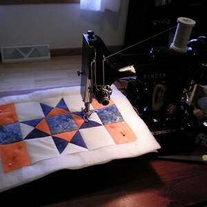 Puede incluir: Una m&aacute;quina de coser Singer negra se est&aacute; utilizando para coser una colcha blanca con cuadrados de tela naranja, azul y blanca. La colcha se est&aacute; cosiendo en una mesa de madera.