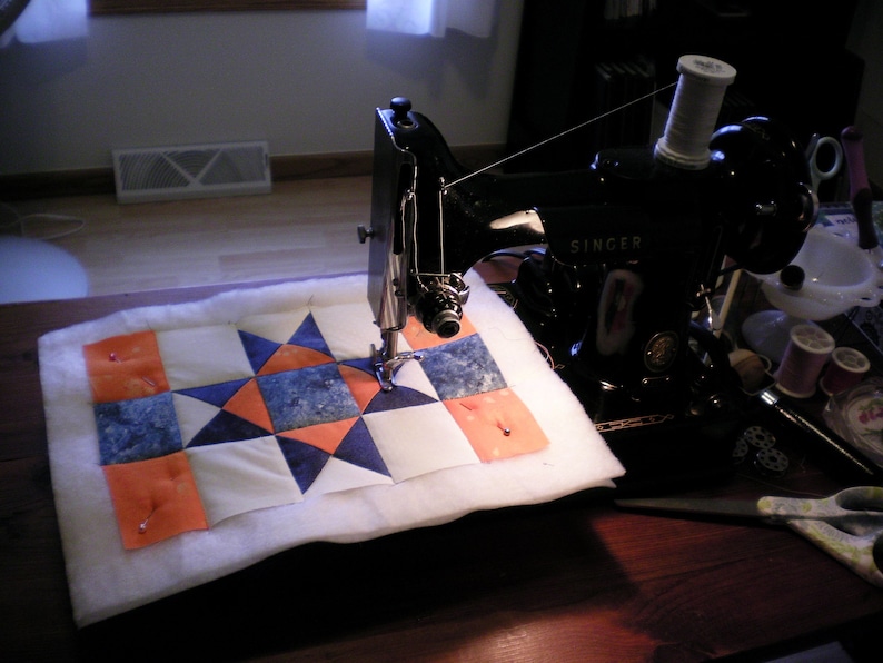 Puede incluir: Una m&aacute;quina de coser Singer negra se est&aacute; utilizando para coser una colcha blanca con cuadrados de tela naranja, azul y blanca. La colcha se est&aacute; cosiendo en una mesa de madera.