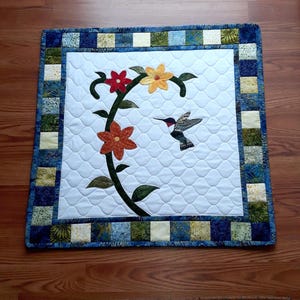 May include: A square quilt with a white centre featuring a floral design and a hummingbird. The flowers are red, orange, and yellow, with green leaves and a black stem. The quilt has a blue and green patterned border.