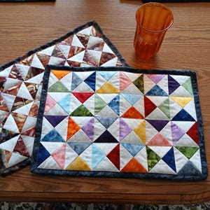 May include: Two handmade quilted placemats with geometric patterns in shades of brown, white, and a variety of colours. A clear orange glass sits on the wooden table. The placemats are rectangular and square.