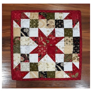 Puede incluir: Un edredón cuadrado hecho a mano con un borde rojo y un diseño de estrella central. La estrella es roja y los cuadrados circundantes presentan varios patrones en blanco, negro, verde y beige. El edredón es probablemente para uso decorativo.