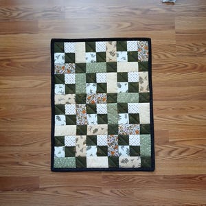 May include: A rectangular quilted wall hanging featuring a patchwork design of alternating solid dark olive green squares and various patterned fabric squares in shades of cream, white, and orange, all bordered by a solid black fabric binding.
