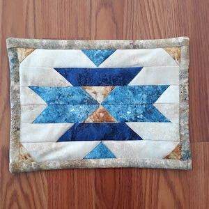 Puede incluir: Un pequeño mantel individual rectangular acolchado con un patrón geométrico en tonos azules, marrones y blancos. El mantel individual tiene un borde marrón.