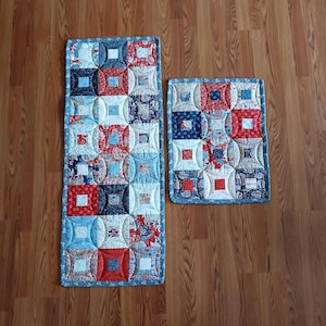 Könnte beinhalten: Zwei gesteppte Tischläufer mit Patchwork-Design. Die Läufer weisen ein Muster aus abwechselnd roten, weißen und blauen Quadraten und Blumendrucken auf. Die Kanten sind mit einem blauen Binding versehen. Einer ist rechteckig, der andere quadratisch.