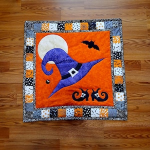 Puede incluir: Una colcha con temática de Halloween con un sombrero de bruja morado, una luna llena, murciélagos y arañas sobre un fondo naranja. La colcha está rodeada por un borde blanco y negro con un patrón de telaraña.