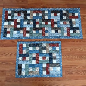 May include: Two handmade quilted table runners. The larger runner is rectangular, and the smaller one is square. Both have a patchwork design with blue, red, white, and brown fabric squares. The edges are finished with a blue floral print.
