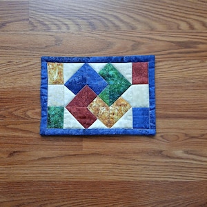 May include: A small, rectangular quilted placemat with a blue border. The centre features a geometric pattern of diamonds and squares in various colours, including blue, green, red, and cream. The placemat is placed on a wooden surface.