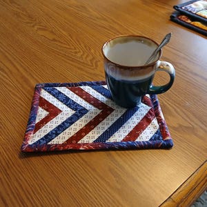 May include: A red, white, and blue quilted coaster with a chevron pattern. A blue and brown mug with a spoon is sitting on the coaster.