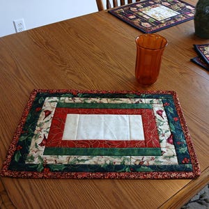 Könnte beinhalten: Ein rechteckiger, gesteppter Tischset mit einem zentralen weißen Feld, umgeben von rotem, grünem und gemustertem Stoff mit Stechpalmen- und Vogelmotiven. Das Tischset liegt auf einem Holztisch, mit einem orangefarbenen Glas und weiteren Tischsets.