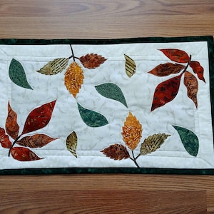 May include: A rectangular quilted table runner with a cream background and a dark green border. The runner features appliqued leaves in shades of red, orange, green, and brown, creating a fall-themed design. The leaves are arranged in a scattered pattern.