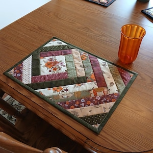 May include: A rectangular quilted placemat featuring a log cabin pattern with various fabric strips in shades of olive green, burgundy, cream, and orange, accented by floral and polka dot prints, accompanied by an orange fluted glass tumbler.