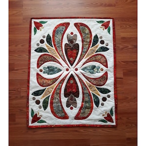 May include: A rectangular quilt with a central floral design. The quilt features a white background with a symmetrical pattern of red, brown, green, and blue leaf-like shapes. The edges are trimmed with a red border, and the quilt is placed on a wooden floor.
